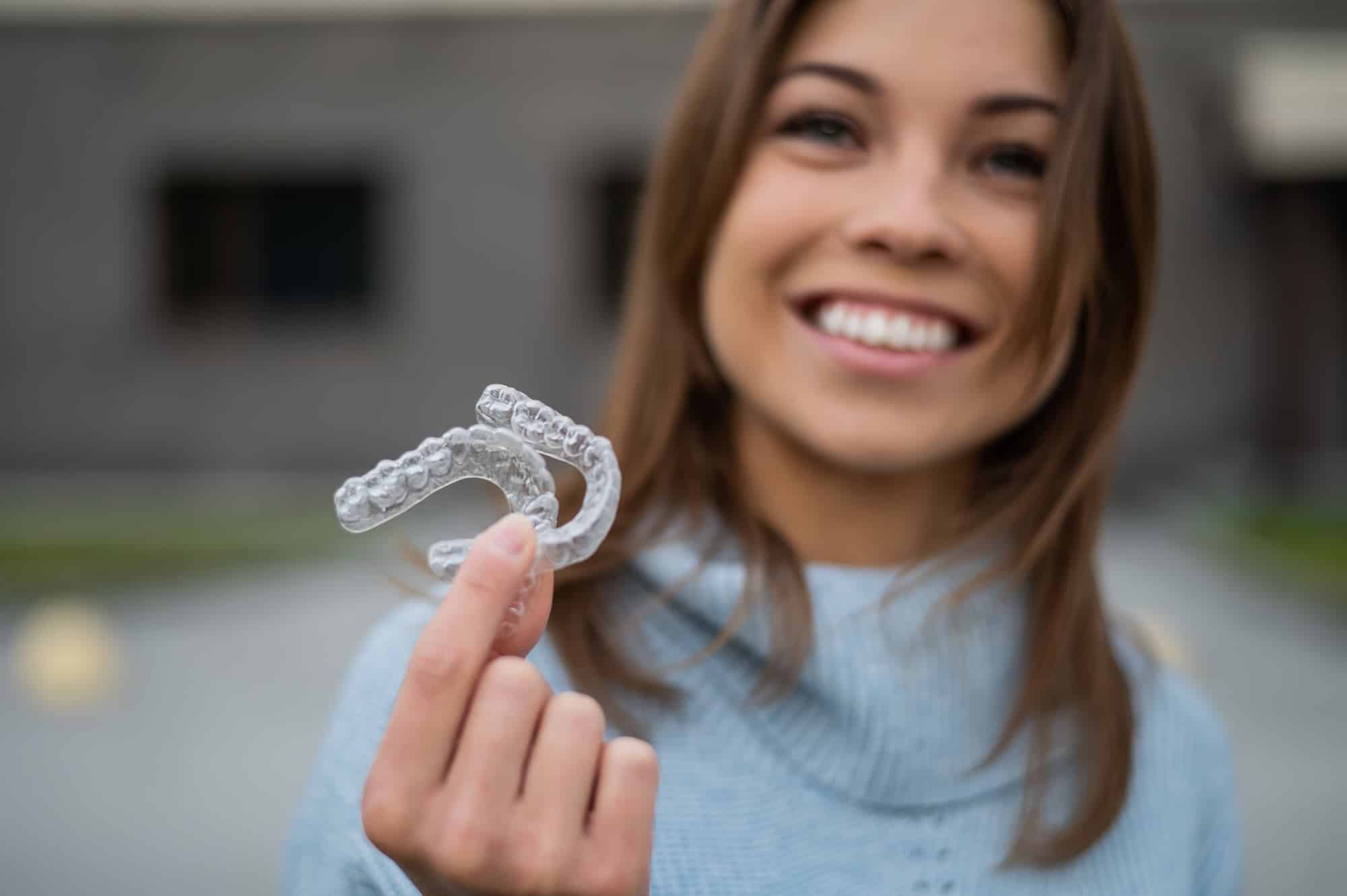 A young woman smiling while holding two transparent dental aligners in her hand. She is wearing a light blue sweater, and the background features blurred buildings and greenery.