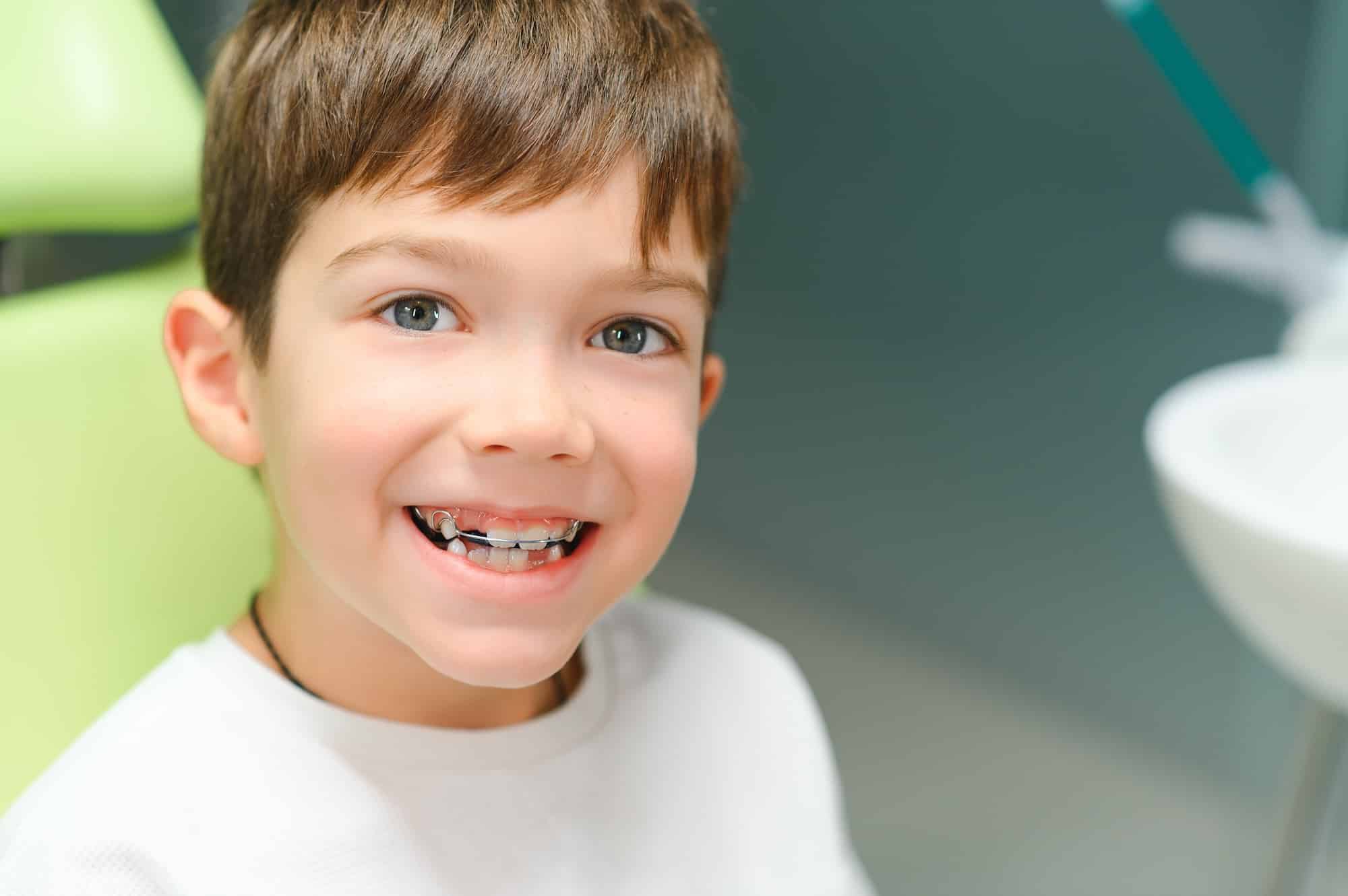A young boy with brown hair smiles while sitting in a dental clinic. He is wearing a white shirt and has dental braces on his teeth. The background shows dental equipment and a green dental chair.