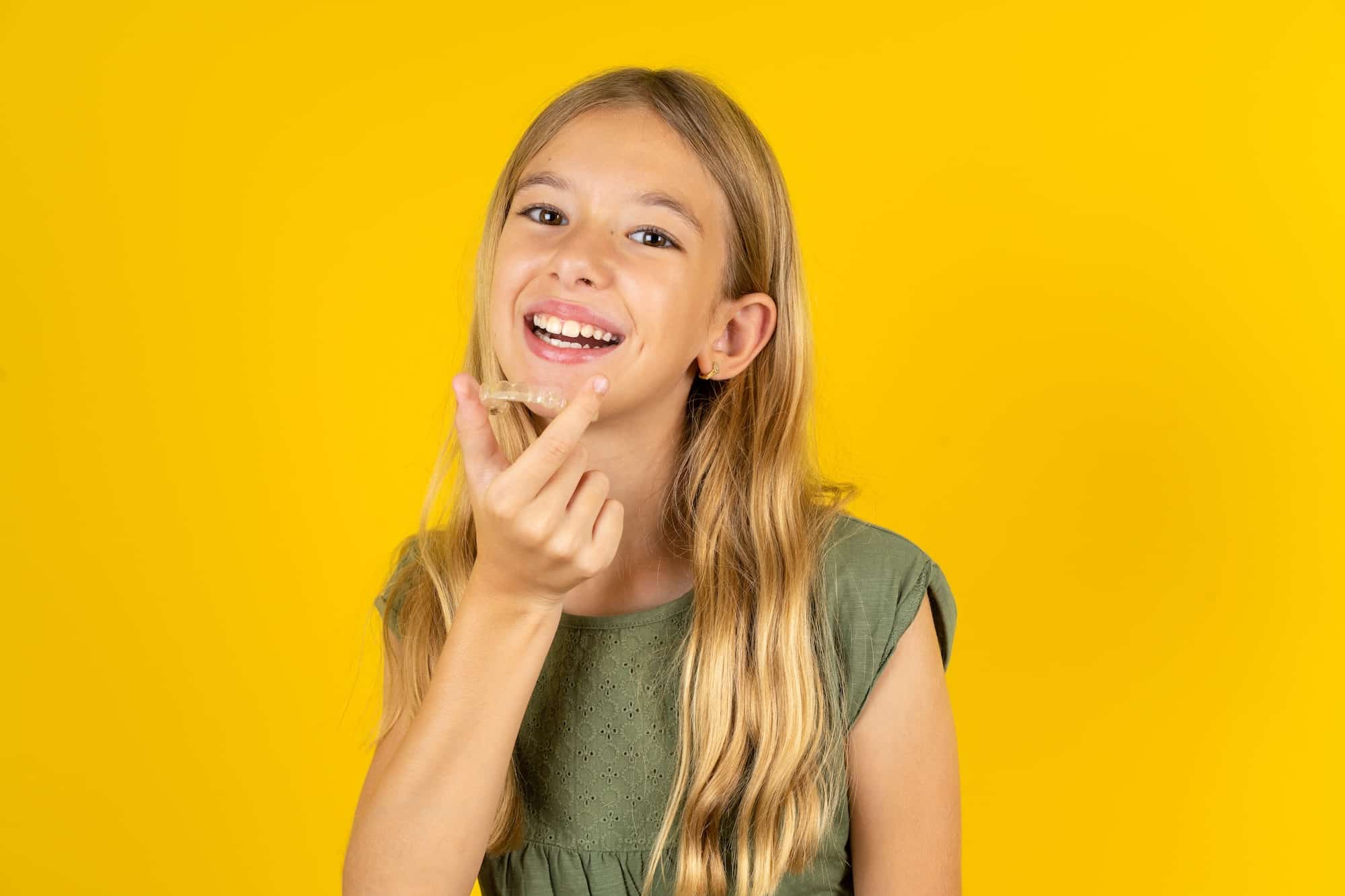 A young girl with long blonde hair smiles while making a hand gesture resembling a heart. She is wearing a green shirt, and the background is a bright yellow, creating a cheerful atmosphere.