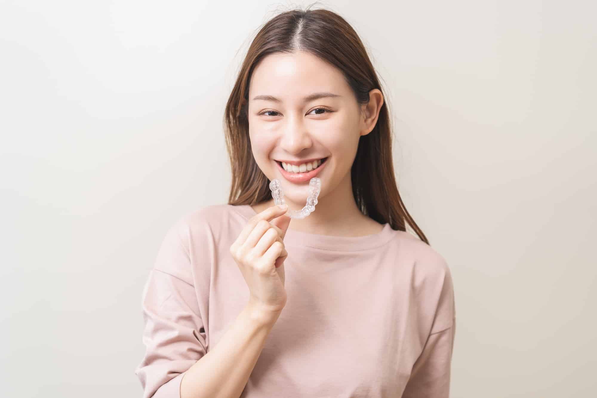 A young woman with long, straight hair smiles while holding an invisible aligner in front of her teeth. She wears a light pink sweater and stands against a neutral background, highlighting her expression and the aligner.