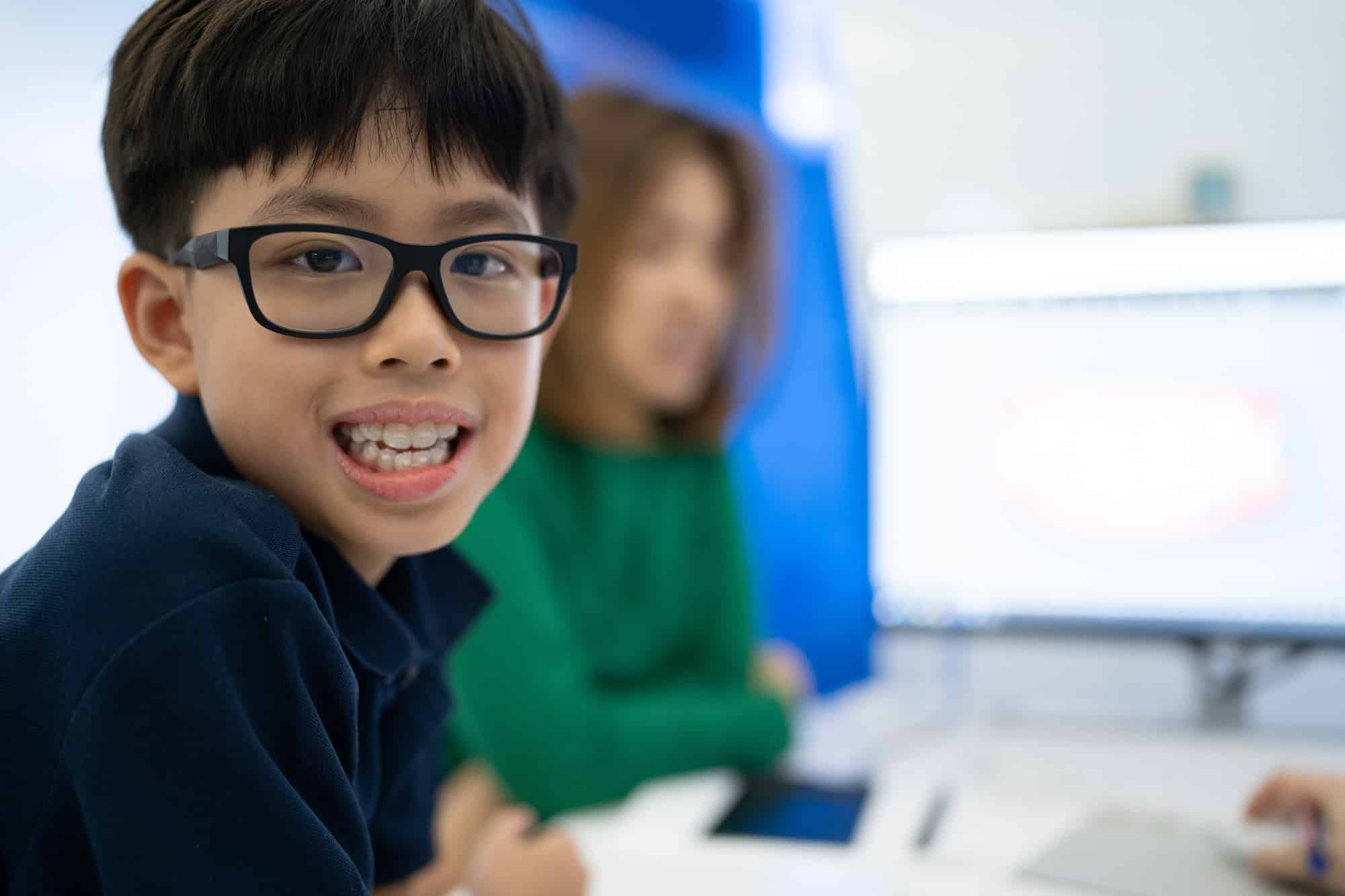 A young boy with glasses smiles at the camera, seated at a table with a computer. A woman in a green shirt is blurred in the background, appearing to engage in a discussion. The setting is bright and modern.