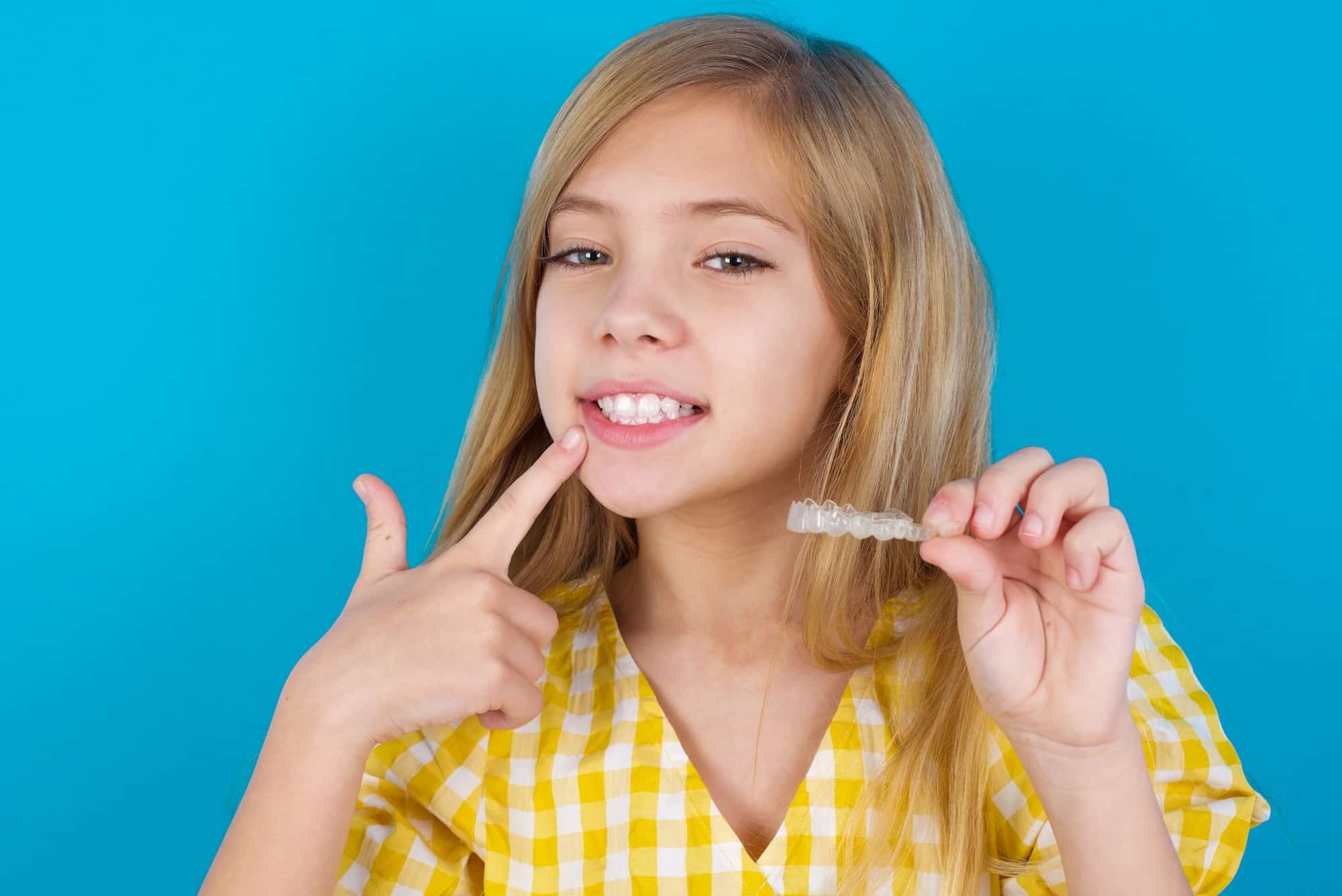 A girl with long blonde hair smiles while holding a clear orthodontic aligner. She is wearing a yellow checkered top and pointing to her teeth with her other hand. The background is a solid blue color.