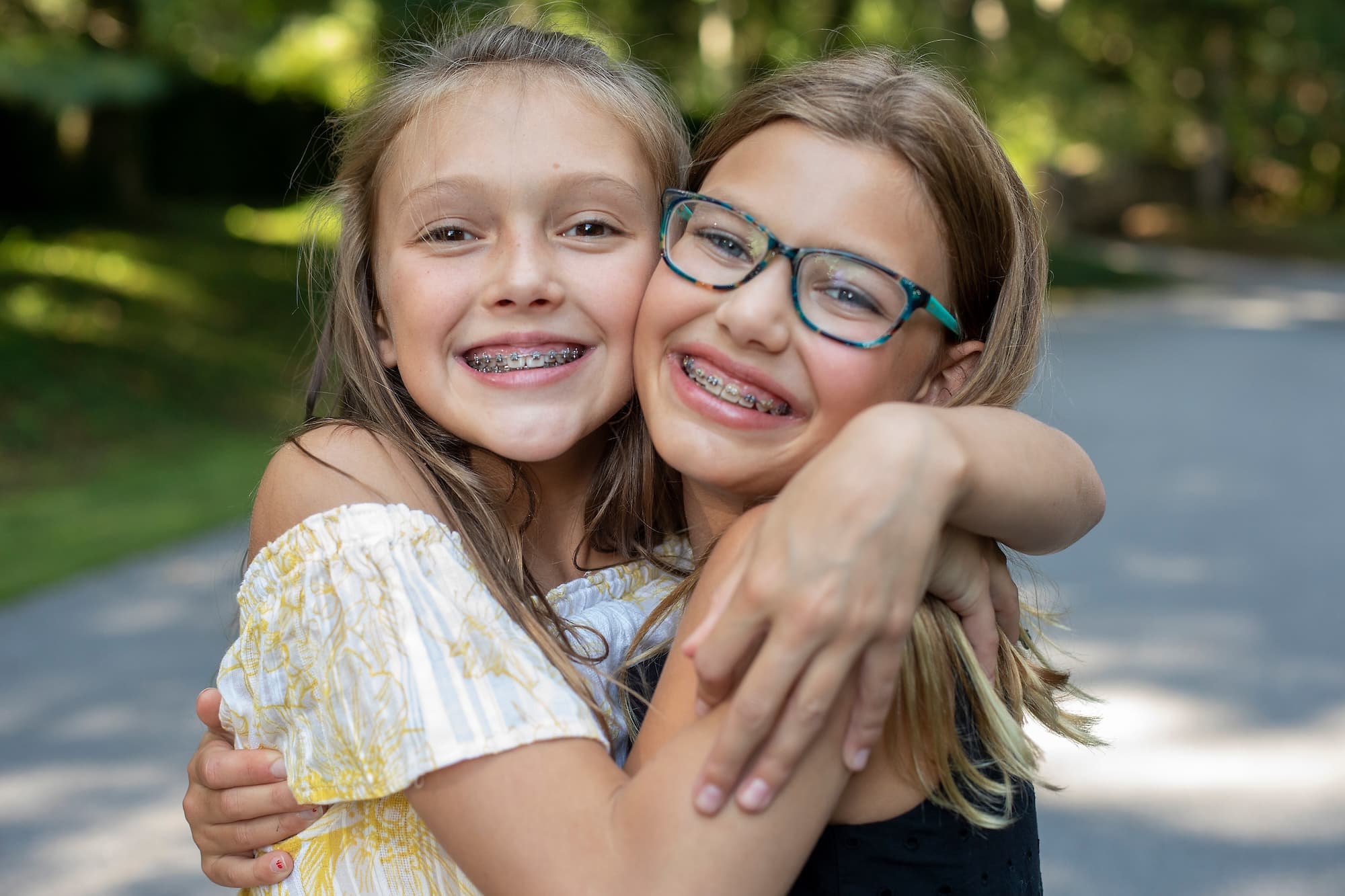 Two girls smiling and embracing each other outdoors. One girl has long brown hair and braces, while the other has medium-length blonde hair and glasses. They are standing on a pathway with greenery in the background.