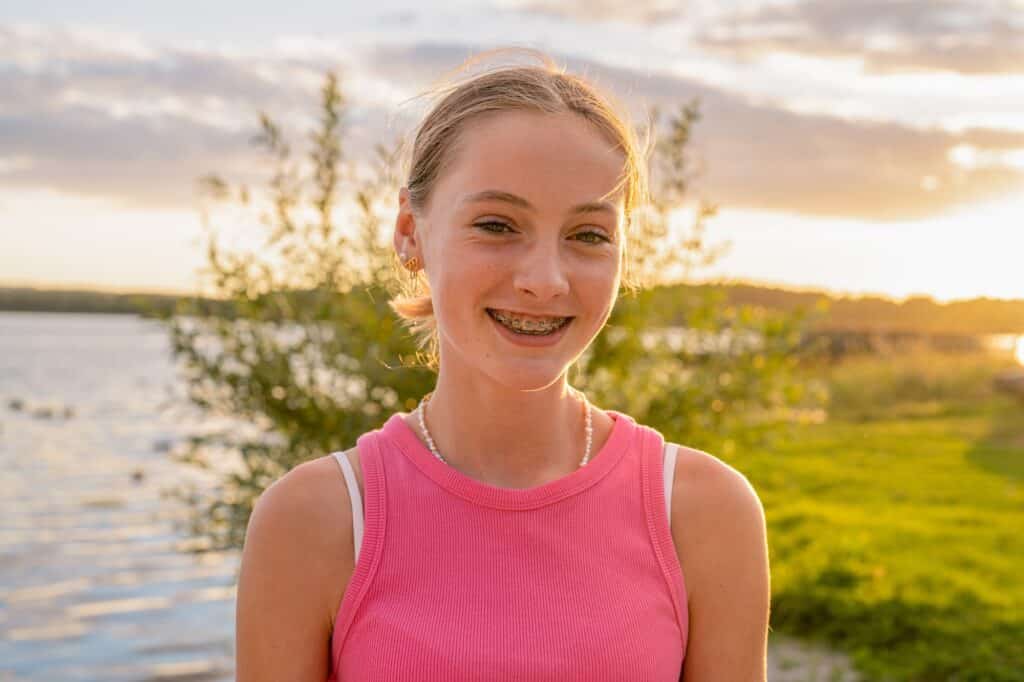 A young girl with braces smiles while standing outdoors near a lake, surrounded by greenery. The sun is setting in the background, casting a warm glow over the scene. She is wearing a pink sleeveless top.