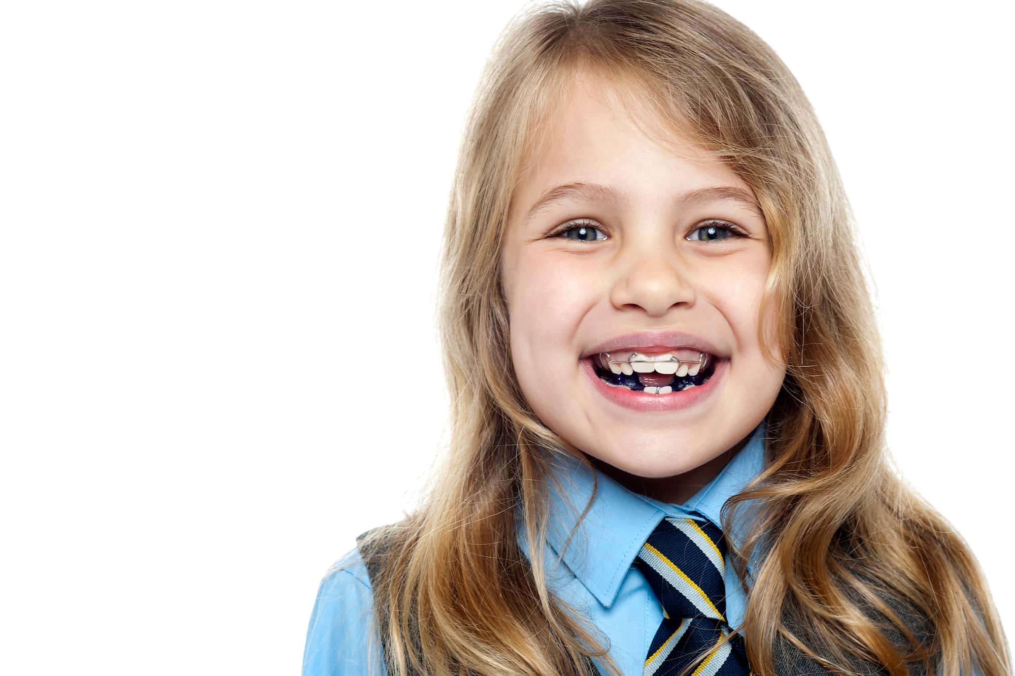 A young girl with long, blonde hair smiles broadly, showing a gap in her front teeth. She wears a light blue shirt and a striped tie, set against a plain white background.