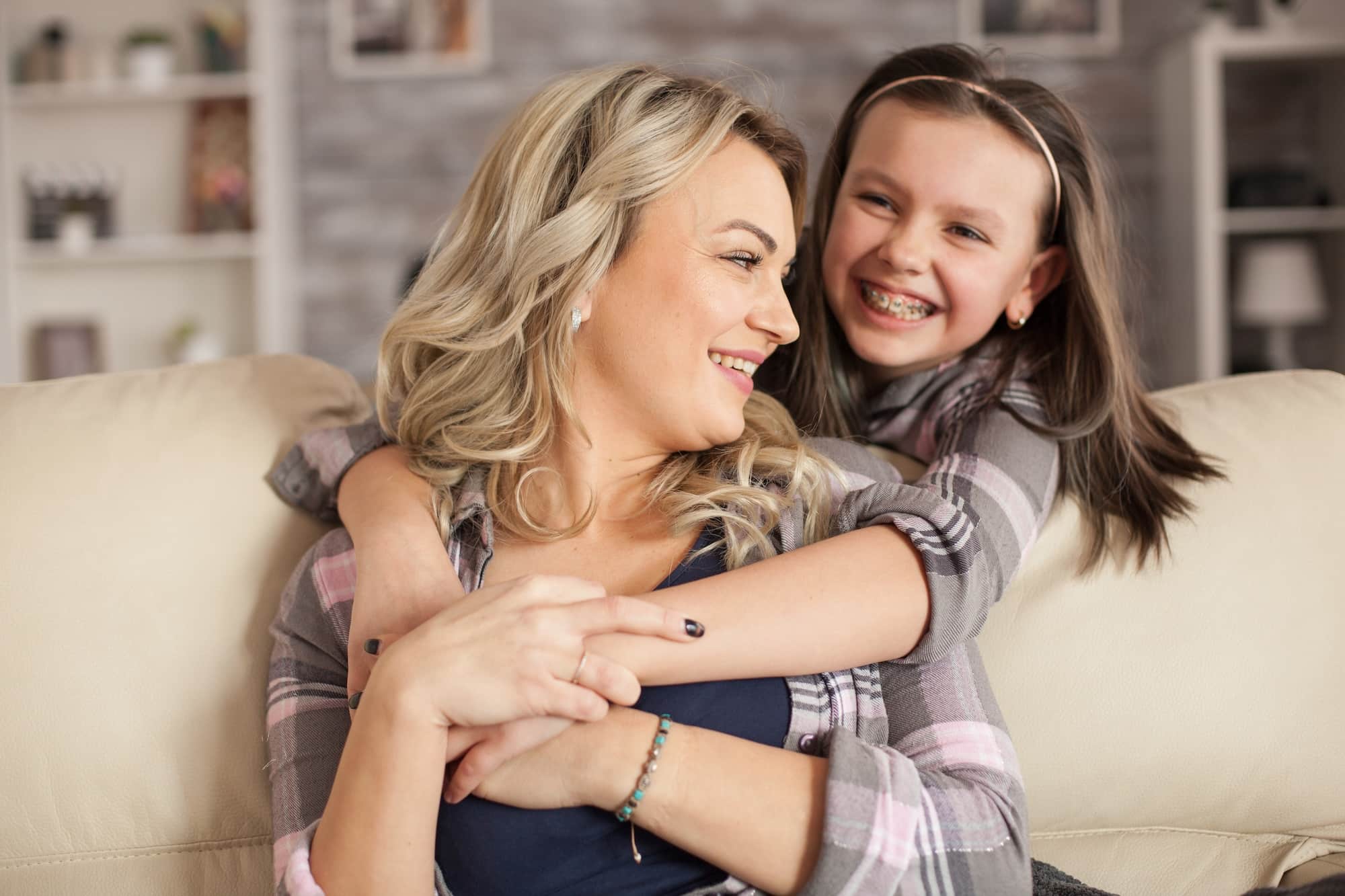 A woman with long, blonde hair is smiling while sitting on a couch, embracing a young girl with dark hair who is grinning widely. Both wear casual outfits, and the background features soft lighting and shelves with decorative items.